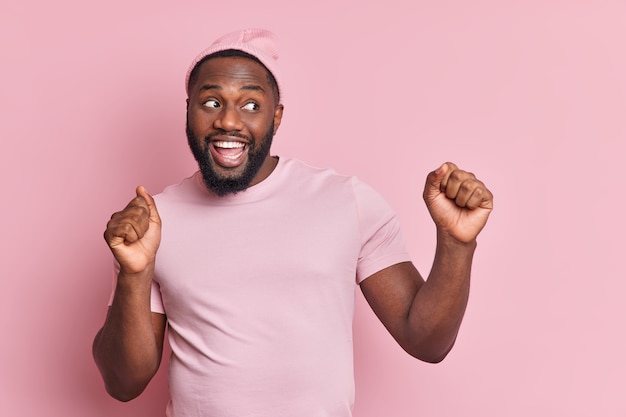 Happy relaxed man dances carefree raises arms clenches fists moves with rhythm of music smiles broadly dressed casually isolated over pink wall