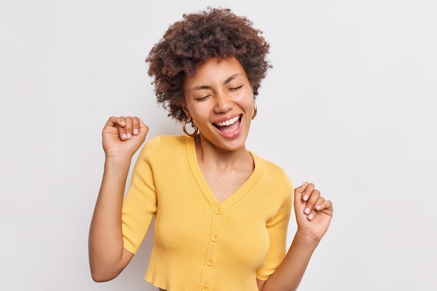 Happy relaxed woman catches carefree moment enjoys freedom sings song keeps arms raised up closes eyes dances to favorite music wears casual yellow t shirt isolated on white wall