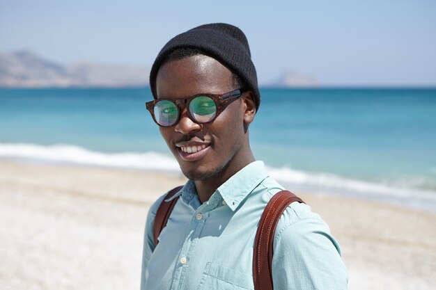 Happy relaxed young dark skinned hipster in shades and headwear standing against clear blue sky and vast azure ocean, spending summer holidays in resort town, looking with joyful smile