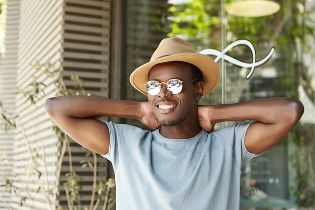 Happy and relaxed young dark-skinned male student in stylish sunglasses and headwear keeping hands behind head and smiling broadly, enjoying summer vacations, having lunch at sidewalk restaurant