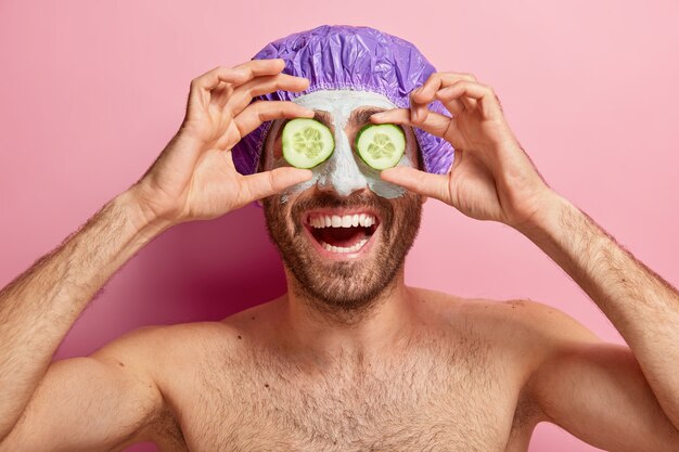 Happy relaxed young man covers eyes with two slices of cucumber, applies cosmetic mask on face, wears bathcap, stands naked against pink wall. Self care, beauty and spa therapy concept