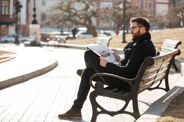 Happy relaxed young man reading newspaper on the bench outdoors