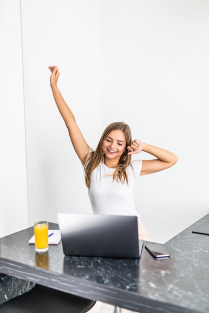Foto gratuita felice e rilassata giovane donna seduta nella sua cucina con un laptop davanti a lei che allunga le braccia sopra la testa