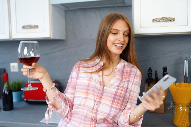레드 와인 한 잔과 함께 부엌에 서서 videocall에 그녀의 스마트 폰을 사용하는 행복 편안한 젊은 여자