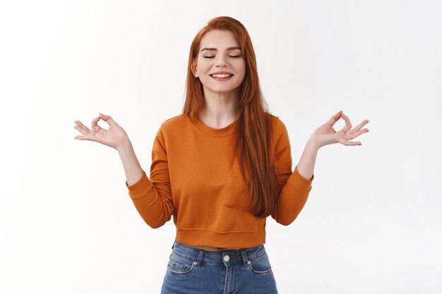Happy relieved calm smiling redhead female close eyes feel relaxation nirvana raise hands zen mudra gesture meditation, exercise yoga breathing relaxing self-soothing practice stand white wall