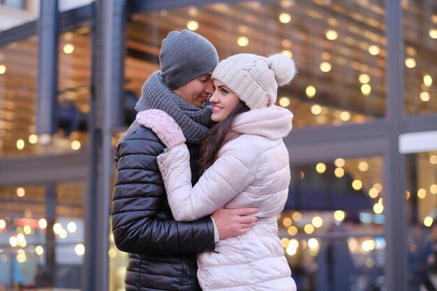 Happy romantic couple wearing warm clothes hugging together in evening street near a cafe outside at Christmas time