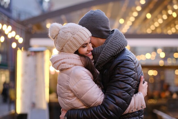 Happy romantic couple wearing warm clothes hugging together in evening street near a cafe outside at Christmas time