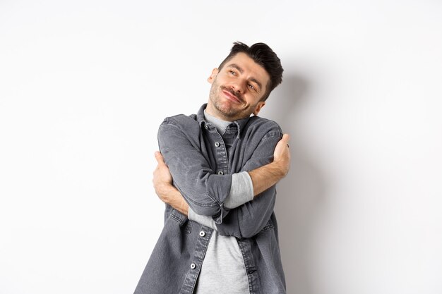 Happy romantic guy hugging himself with closed eyes and smile, loving own body, daydreaming about lover, standing on white background.