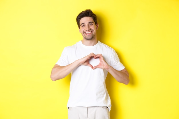 Felice uomo romantico che mostra il segno del cuore, sorride ed esprime amore, in piedi su sfondo giallo