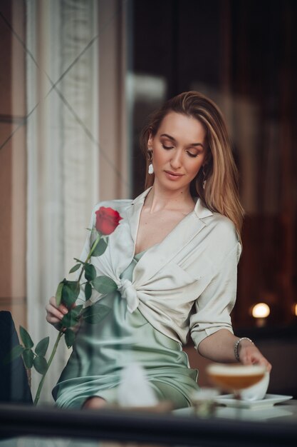 Foto gratuita felice giovane e bella ragazza romantica con un fiore di rosa rossa che sogna seduta al bar durante gli appuntamenti del giorno di san valentino