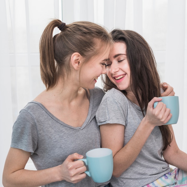 Happy romantic young lesbian couple in grey t-shirt holding blue cup of coffee in hand