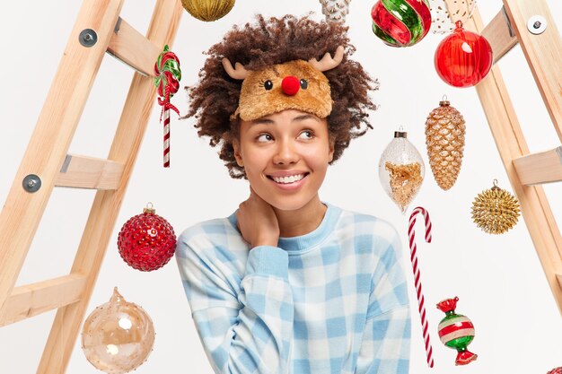 Счастливая романтичная молодая женщина с вьющимися волосами ждет счастливого Рождества, наслаждается уютной домашней атмосферой, носит маску для сна из оленей, а в пижаме используется лестница, чтобы вешать игрушки на елку. Концепция зимнего времени