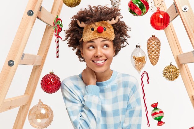 Happy romantic young woman with curly hair waits for Merry Christmas enjoys cozy domestic atmosphere wears reindeer sleepmask and pajama uses ladder to hang toys on fir tree. Winter time concept