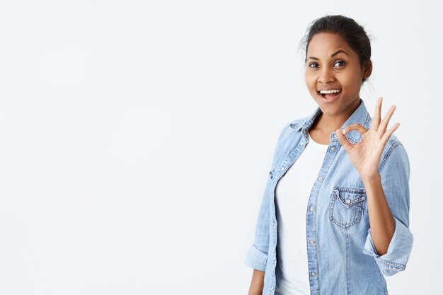 Happy satisfied Afro-american female wearing denim shirt making okay sign and smiling cheerfully, showing her support and respect to someone. Positive emotions concept.