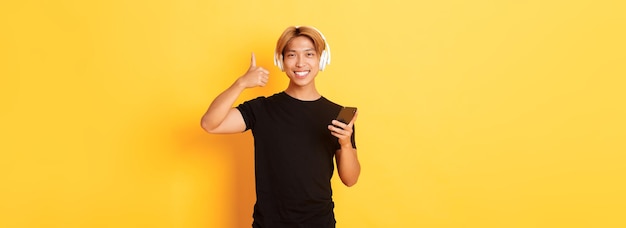 Happy satisfied asian guy likes music or podcast showing thumbsup in approval holding mobile phone s