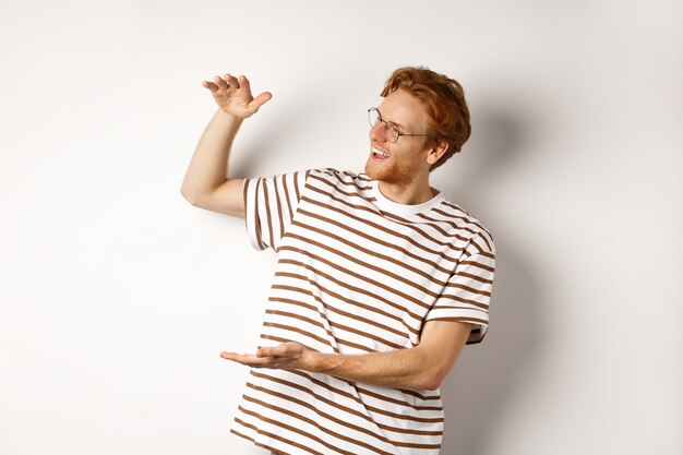 Happy and satisfied caucasian redhead man introduce something big, showing large product and smiling pleased, white background