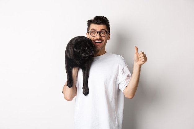 Happy and satisfied dog owner showing thumb-up, holding cute black pug on shoulder, recommending pet products, standing over white background