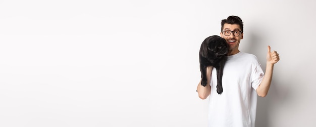 Free photo happy and satisfied dog owner showing thumbup holding cute black pug on shoulder recommending pet pr