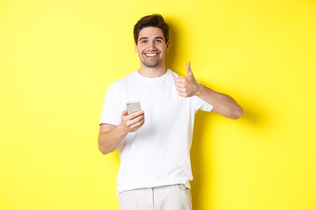 Happy satisfied man holding smartphone, showing thumb up in approval, recommend something online, standing over yellow background