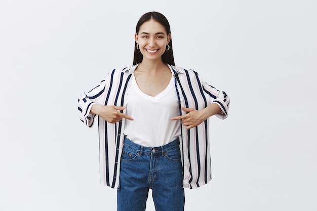 Happy satisfied woman standing in trendy outfit and pointing at belly, showing she lost weight, bragging about results after visiting gym, standing healthy and sporty over gray wall