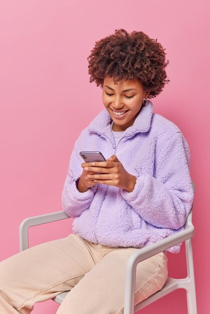 Happy satisfied woman with curly bushy hair checks notification via smartphone wears purple fur coat and trousers poses on comfortable chair smiles gladfully reads text message from boyfriend