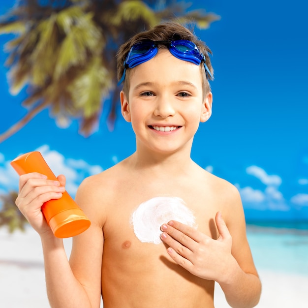 Happy schoolchild boy applying sun block cream on the tanned body. boy holding orange sun tan lotion bottle.