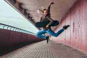 Foto gratuita una studentessa felice con i capelli biondi, vestita con abiti alla moda, salta con uno skateboard sul marciapiede del ponte.