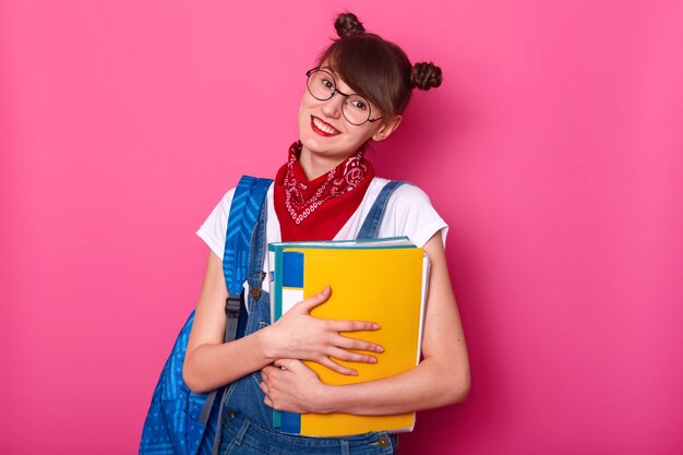 バラ色に分離された紙フォルダーと幸せな女子高生。夏の休日の後に学校に戻って喜んでいる笑顔の女の子。女性はTシャツとオーバーオールを着て、頭を傾けて笑顔