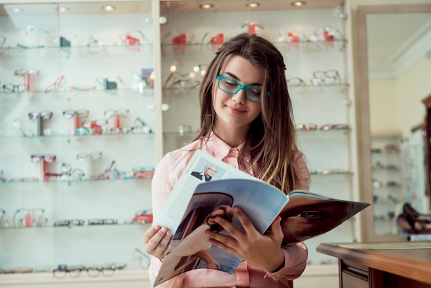 Free photo happy to see words clearly without blur. indoor portrait of satisfied attractive european woman sitting in optician store while reading magazine in glasses, waiting for het turn to check sight