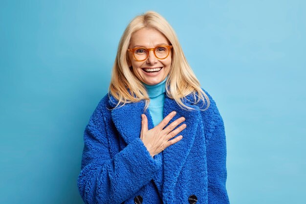happy senior blonde European woman amused by humorous joke laughs positively keeps hand on chest dressed in winter blue coat.