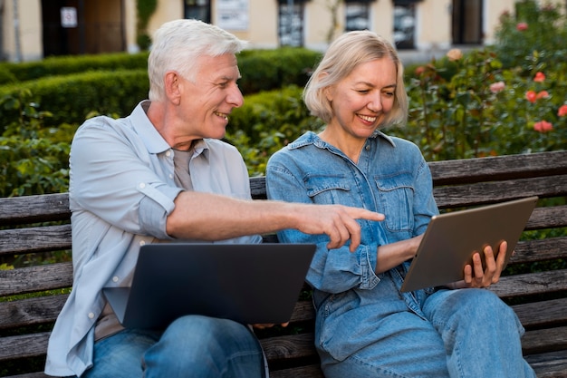 Happy senior couple on bench outdoors with laptop and tablet