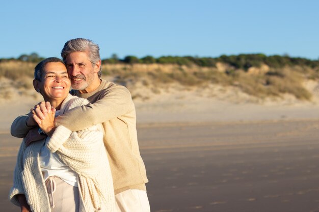 Happy senior couple having fun at seashore hugging and smiling. Bearded man embracing lady with short hair, intertwining fingers. Romance, leisure, retirement concept