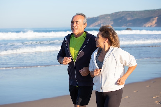 夏の日に海の海岸に沿って走っている幸せな年配のカップル。有酸素運動トレーニングを受けている、健康管理をしている白人男性と女性の笑顔のミディアムショット。家族、スポーツの概念