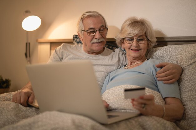 Happy senior couple shopping on the Internet with laptop and credit card while lying down in bed in the evening