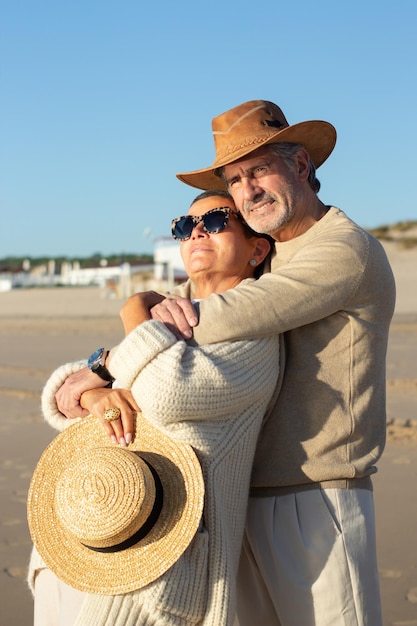 Free photo happy senior couple standing at seashore enjoying sea view on warm sunny autumn evening. short-haired lady in sunglasses holding straw hat and leaning on husbands chest. relationship, love concept