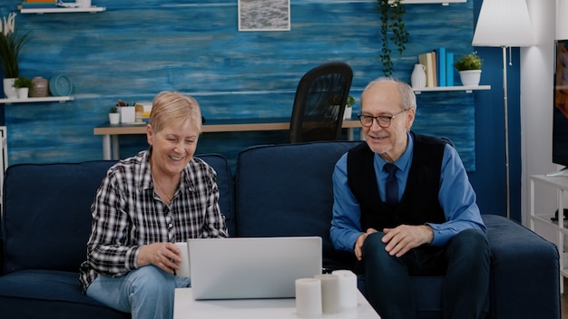 Happy senior couple waving at videocall with nephews using laptop sitting on couch in living room