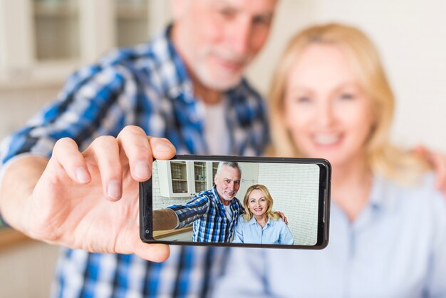 Happy senior husband and wife are making selfie on mobile phone in kitchen