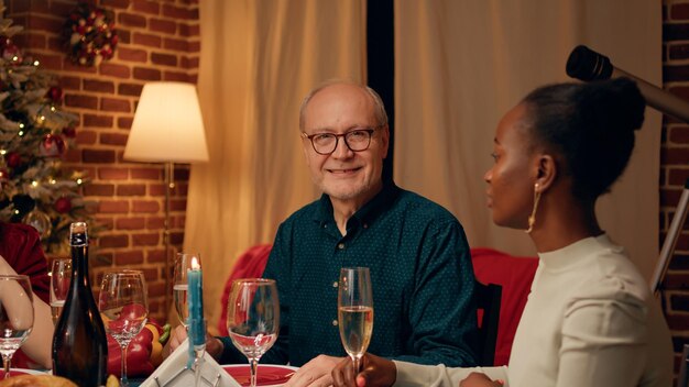 Happy senior man enjoying Christmas dinner table together with close family members while smiling heartily at camera. Joyful person sitting in living room while celebrating traditional winter holiday.