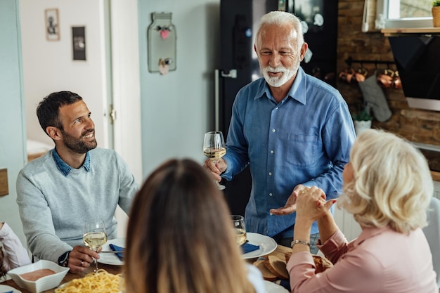 Счастливый пожилой мужчина держит бокал вина, предлагая тост во время семейного обеда