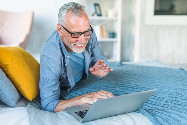 Happy senior man lying on bed using laptop at home