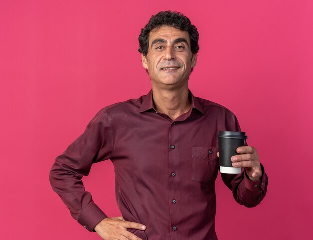 Happy senior man in purple shirt holding paper cup looking at camera smiling confident 