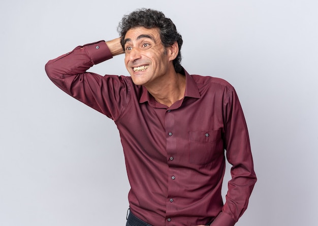 Happy senior man in purple shirt looking aside amazed and surprised with hand on his head standing over white background