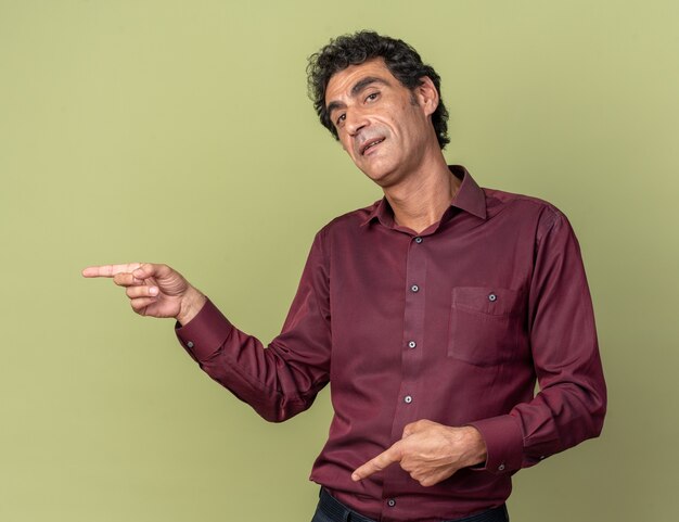 Happy senior man in purple shirt looking at camera smiling pointing with index fingers to the side standing over green