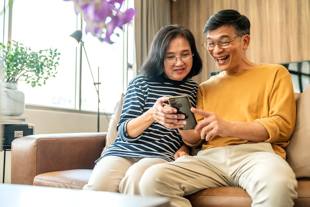 스마트폰을 들고 휴대폰 화면을 보고 있는 행복한 노년의 아시아 연인 부부는 소파에 캐주얼하게 앉아 웃고 있는 노년의 조부모 가족이 생활 방식을 껴안고 웃고 있습니다.