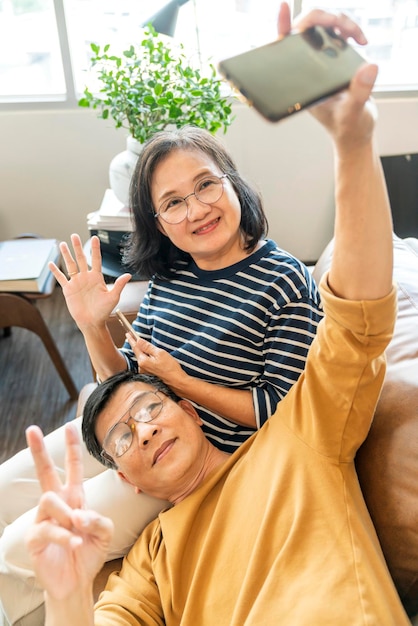 스마트폰을 들고 휴대폰 화면을 보고 있는 행복한 노년의 아시아 연인 부부는 소파에 캐주얼하게 앉아 웃고 있는 노년의 조부모 가족이 생활 방식을 껴안고 웃고 있습니다.