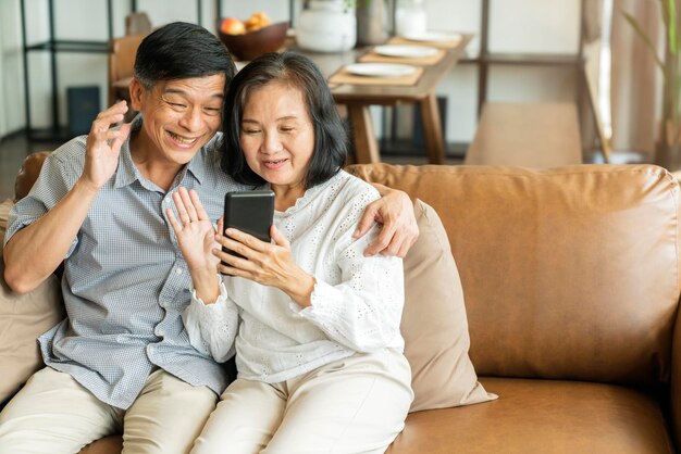 スマートフォンを持って携帯電話の画面を見て、カジュアルにリラックスして笑っている幸せなシニアの古いアジアの恋人のカップルが一緒にソファに座って笑顔の高齢者の成熟した祖父母の家族のライフスタイルを受け入れる