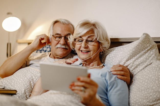 Happy senior woman and her husband using digital tablet while relaxing in bed at home