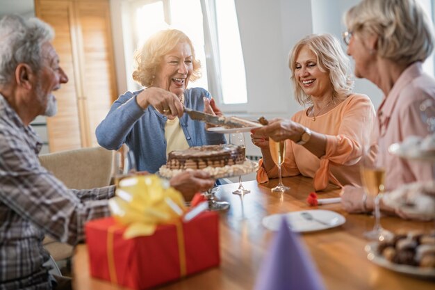 Счастливая пожилая женщина подает торт своим друзьям во время празднования дня рождения дома