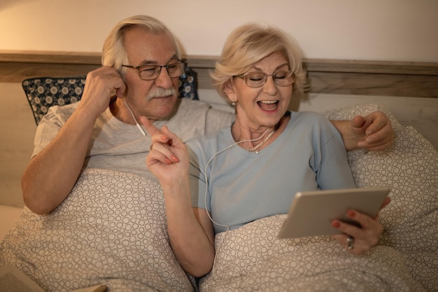 Happy senior woman signing while listening music with her husband on touchpad in bedroom