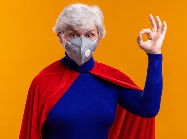 Happy senior woman superhero wearing red cape and facial protective mask looking at camera showing ok sign standing over orange background